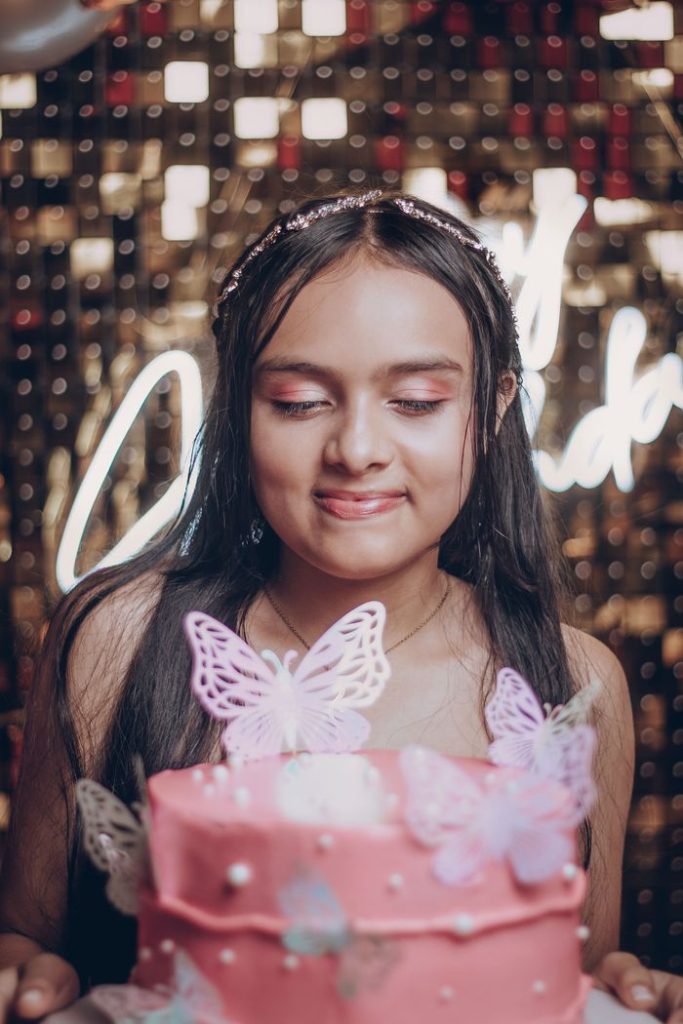 Smiling girl celebrates her birthday with a pink butterfly cake indoors.